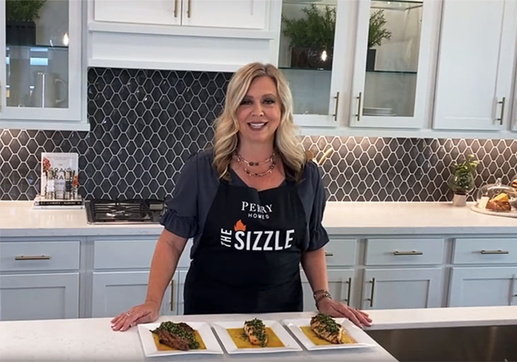 Woman stands behind kitchen island that has three plates of food topped with green Chimichurri sauce.