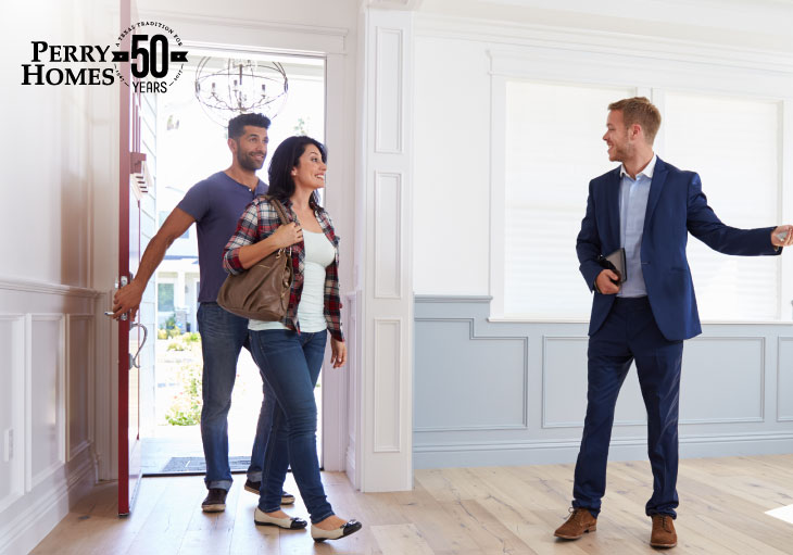 couple walk through red front door to see real estate agent in empty room with white and grey walls and light wood floors