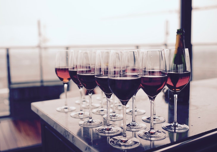 fifteen wine glasses filled with red wine on bar with wine bottle next to them and bright blurred background