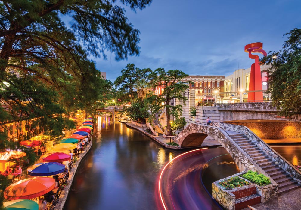 view from street level of san antonio riverwalk at dusk with colorful dining table umbrellsas and partly cloudy sky
