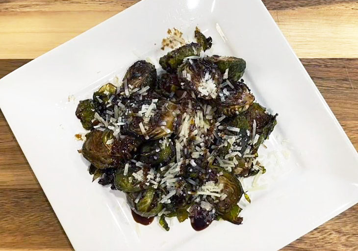 Photo of roasted Brussels sprouts on white rectangular dish with freshly grated parmesan.