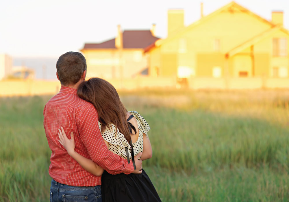 view from behind of young couple embracing as they look at homes in the distance while standing in tall grass field at dusk