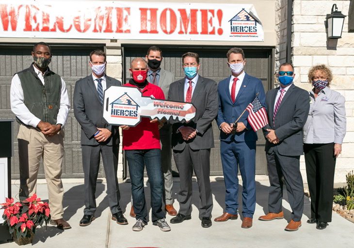 Group of people holding signs in the shape of keys, stand in front of new home with a welcome home sign.