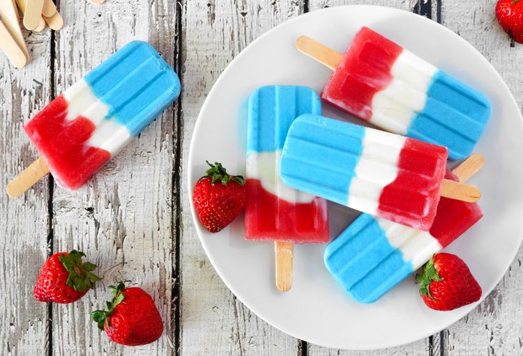 A group of red, white and blue popsicles is laid out on a driftwood table surrounded by ripe strawberries.